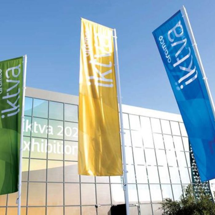Three vertical flags in green, yellow, and blue stand outside a modern building with glass windows and a welcome sign for the "iktva 2023 Forum & Exhibition.