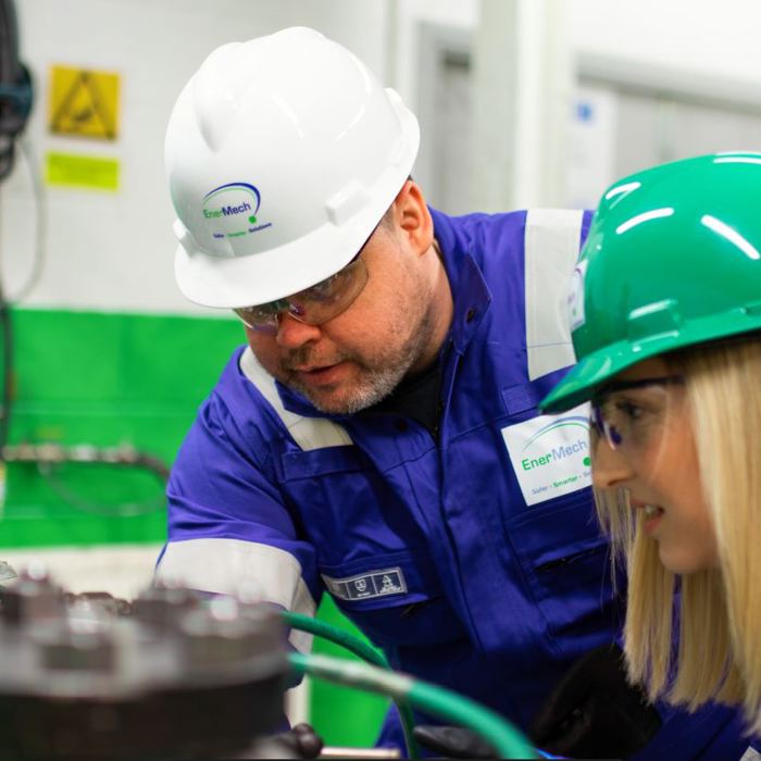Two workers wearing hard hats and protective clothing inspect machinery in an industrial setting. One worker points out a detail to the other.