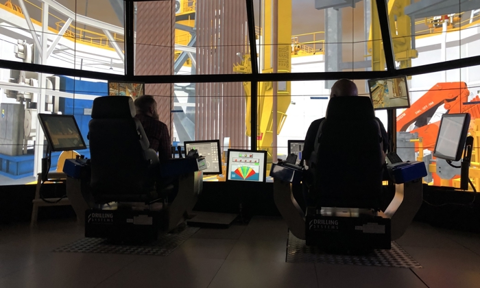 Two individuals operate computer systems in a control room with multiple monitors, overseeing heavy industrial machinery in the background.