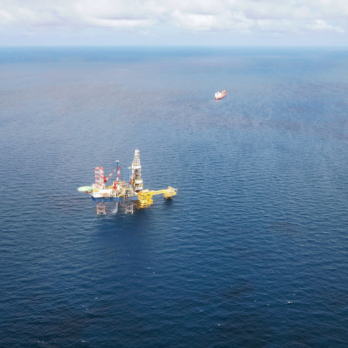 Aerial view of an offshore oil platform in the ocean, with a distant ship on the horizon under a partly cloudy sky, subtly illustrating nature's delicate balance amid the relentless training of industry.