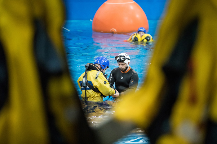 En el centro de una piscina, rodeados de otras personas con trajes amarillos, se ve a dos individuos con equipo para actividades acuáticas, con una gran boya naranja al fondo.