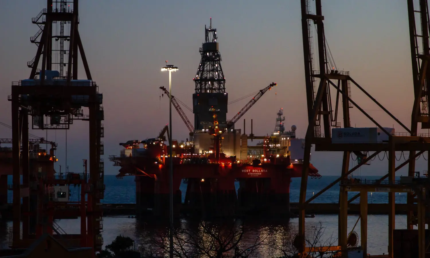 Una plataforma petrolífera en alta mar iluminada al atardecer, rodeada por las siluetas de grandes grúas y estructuras industriales, con el mar de fondo.