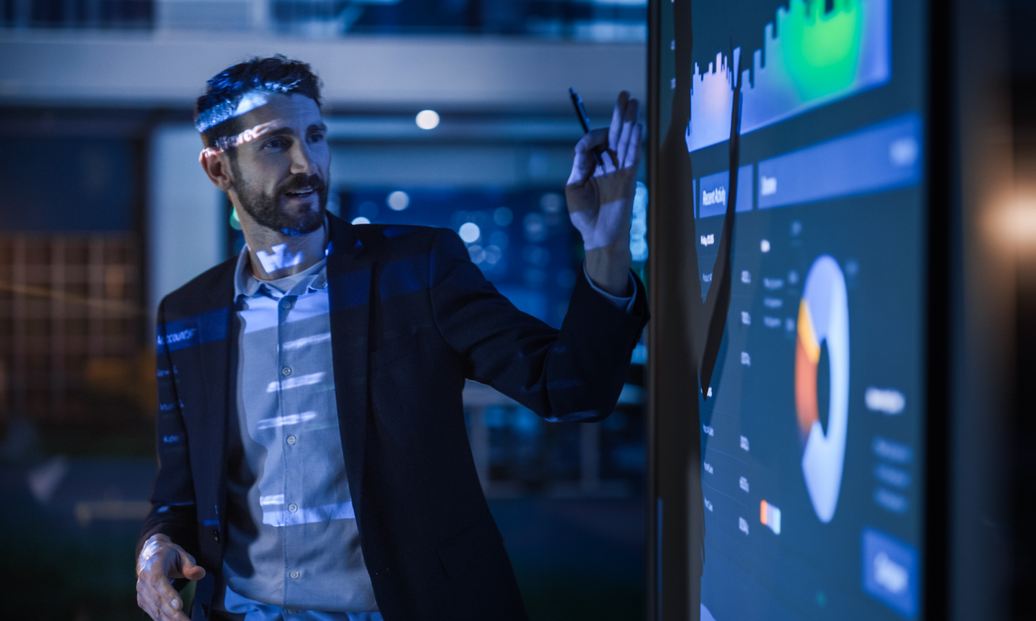 Un hombre con traje de negocios señala una gran pantalla interactiva en la que se muestran gráficos y diagramas en una oficina moderna.