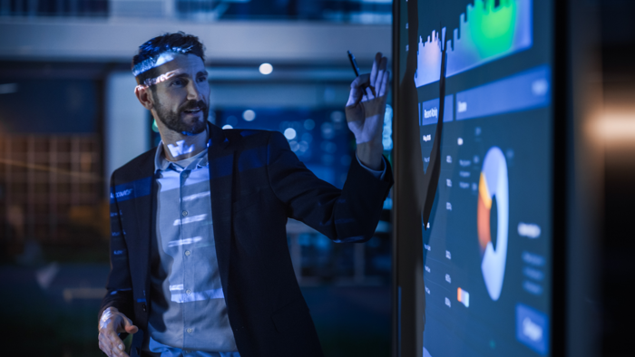 Un hombre con traje de negocios señala una gran pantalla interactiva en la que se muestran gráficos y diagramas en una oficina moderna.