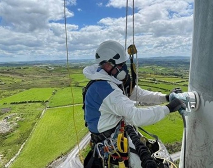 Una persona con equipo de seguridad y casco trabaja en una estructura en lo alto de un paisaje rural con campos verdes bajo un cielo parcialmente nublado.