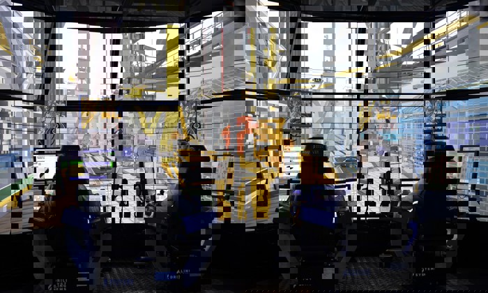 Two people sit at control stations with multiple monitors, operating a high-tech drilling simulator. Large screens simulate an offshore drilling environment around them.