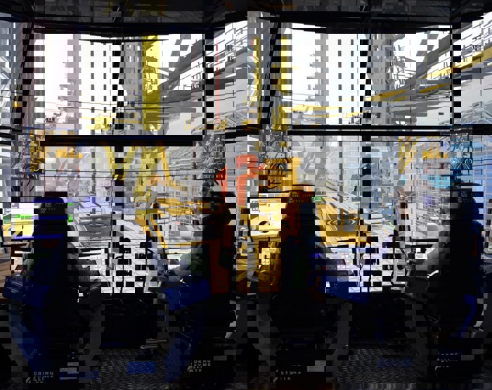 Two people sit at control stations with multiple monitors, operating a high-tech drilling simulator. Large screens simulate an offshore drilling environment around them.