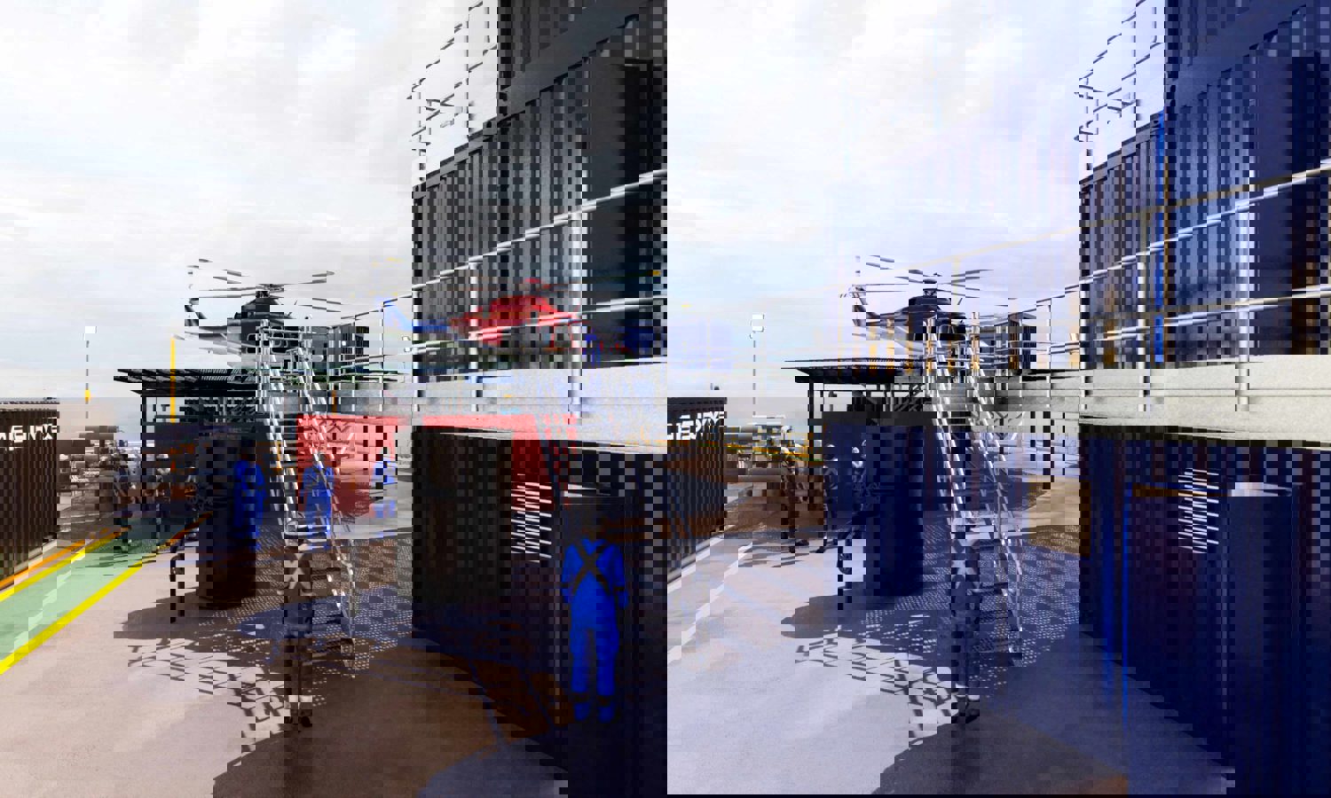 Helicopter on a platform with workers in blue uniforms nearby, situated beside metal containers.