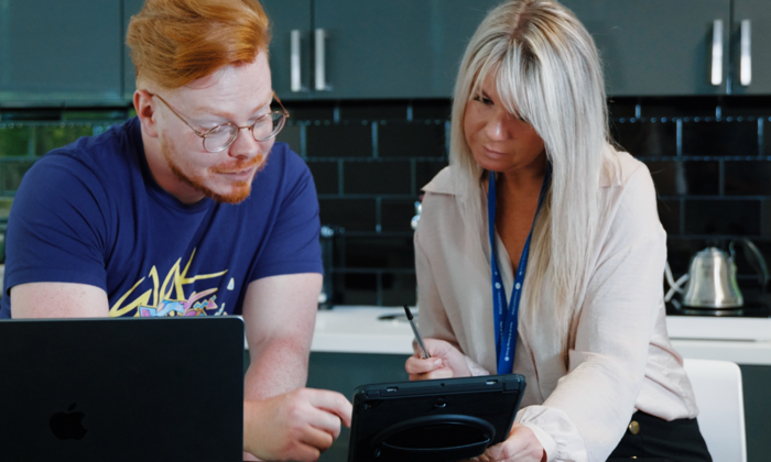 Two people collaborating at a desk, engaged in training. One is holding a tablet, while the other types on a laptop, discussing essential details displayed on the screen.
