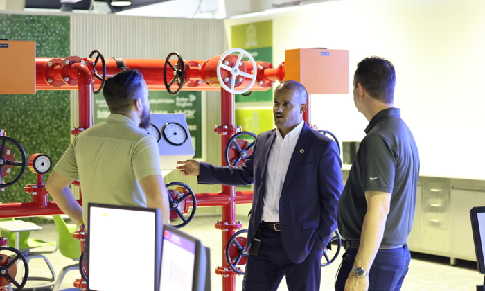 Three men engaged in a training session stand in front of a red industrial pipe setup with gauges and valves, discussing its intricate details.