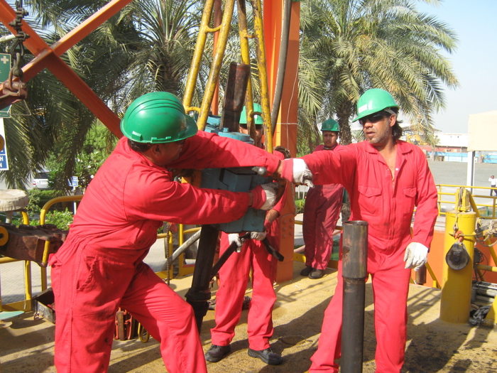 Workers in red coveralls and green helmets operate machinery on an oil rig, surrounded by palm trees.
