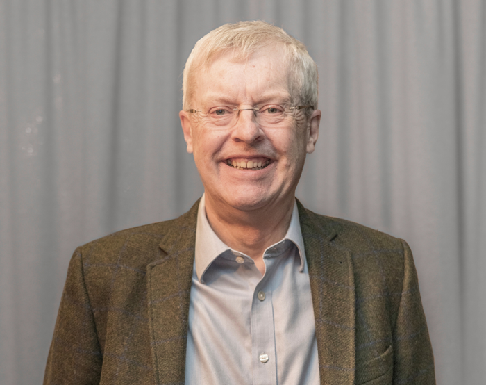 Un hombre mayor con gafas y chaqueta gris sonríe con confianza, posiblemente reflexionando sobre años de entrenamiento, delante de una cortina gris.