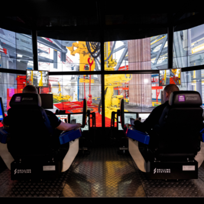 Two people seated in simulator chairs facing multiple screens displaying a construction site environment, with control panels in front of them.