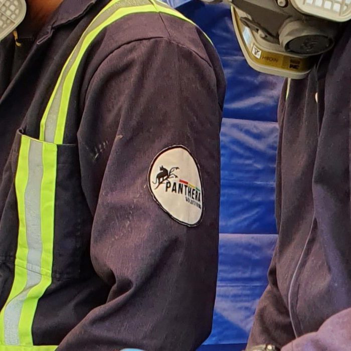 Three individuals wearing protective gear, including face masks and gloves, working closely together. Their dark blue uniforms feature bright green safety stripes and a patch reading "Panthera Solutions.