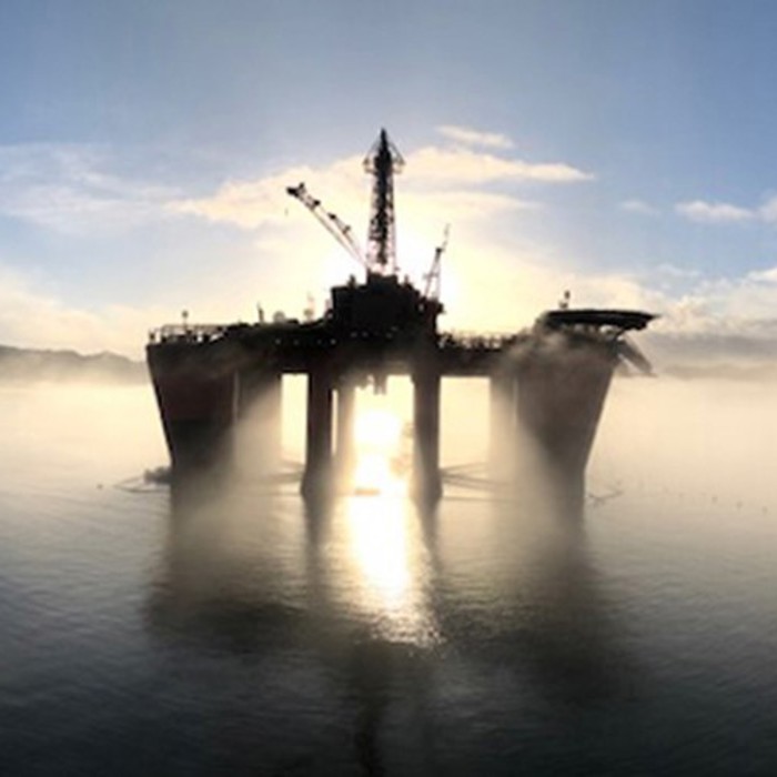 An offshore oil platform stands in the middle of the water with the sun setting behind it, casting a silhouette and reflecting on the water's surface. Mist surrounds the platform.