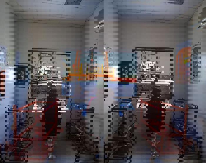 Interior de una sala de control con una persona sentada ante una consola, vigilando una grúa que eleva un contenedor en el exterior. La sala está flanqueada por bastidores rojos y en las paredes hay carteles informativos.