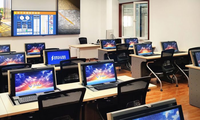 A computer lab with multiple rows of desks, each equipped with a desktop computer featuring brightly colored screensavers. The room includes a large display screen at the front.