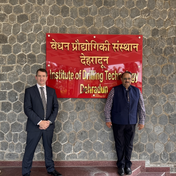 Two individuals stand in front of a red sign with text in Hindi and English that reads "Institute of Drilling Technology Dehradun," attached to a stone wall.