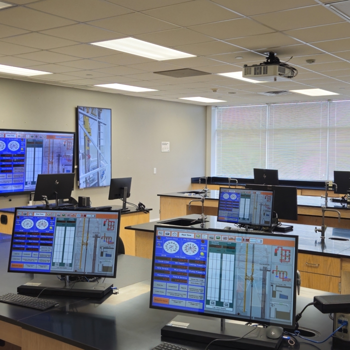 A modern control room with multiple workstations, each featuring monitors displaying control system interfaces. Large wall-mounted screens show surveillance camera feeds.