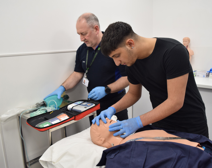 Dos personas practican la reanimación cardiopulmonar en un maniquí médico. Una monitoriza el equipo, mientras la otra realiza compresiones torácicas. Ambos llevan guantes.