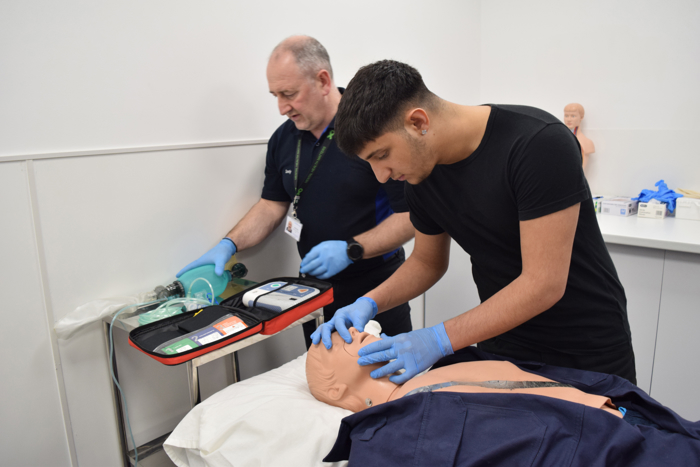 Dos personas practican la reanimación cardiopulmonar en un maniquí médico. Una monitoriza el equipo, mientras la otra realiza compresiones torácicas. Ambos llevan guantes.