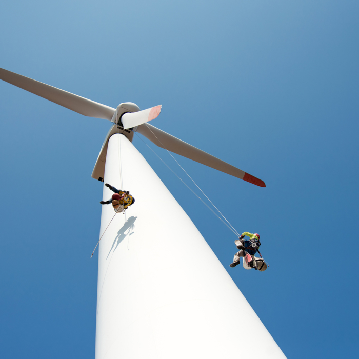 Two workers, supported by ropes, carry out maintenance on a large white wind turbine against a clear blue sky. Their expertise is the result of funded training programs designed to ensure safety and efficiency in this high-risk environment.