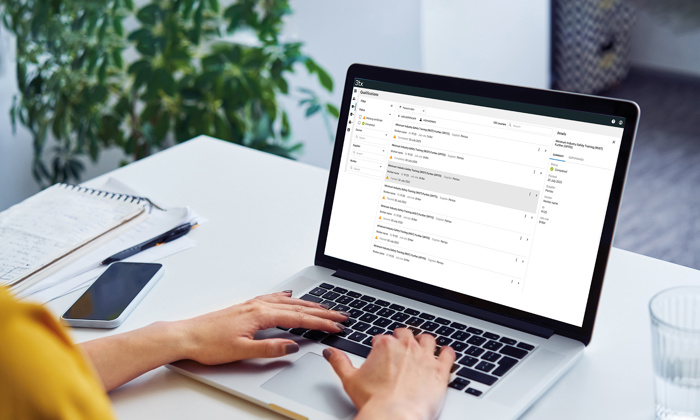 Person typing on a laptop at a desk with an open email inbox on the screen, next to a notebook, pen, smartphone, and a glass of water. A plant is in the background.