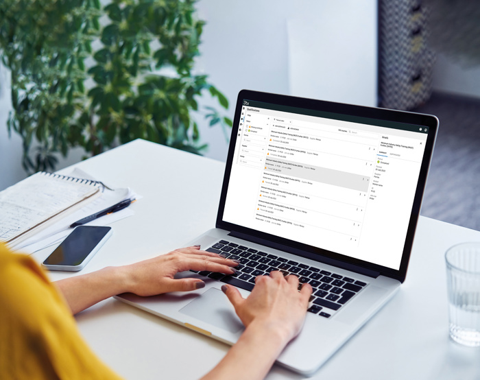 Person typing on a laptop at a desk with an open email inbox on the screen, next to a notebook, pen, smartphone, and a glass of water. A plant is in the background.