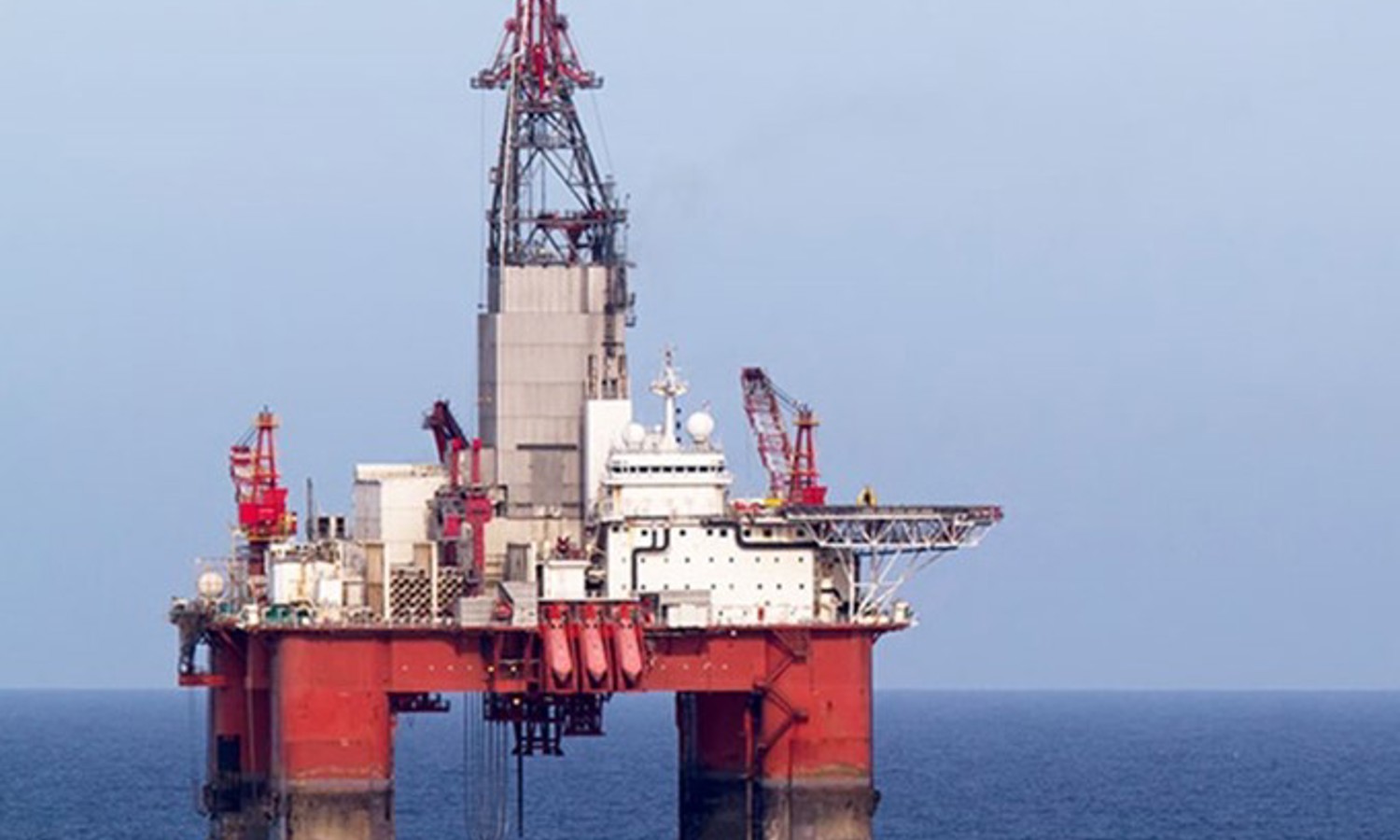 An offshore oil rig stationed in the ocean, with red and white machinery and equipment visible against a clear sky.