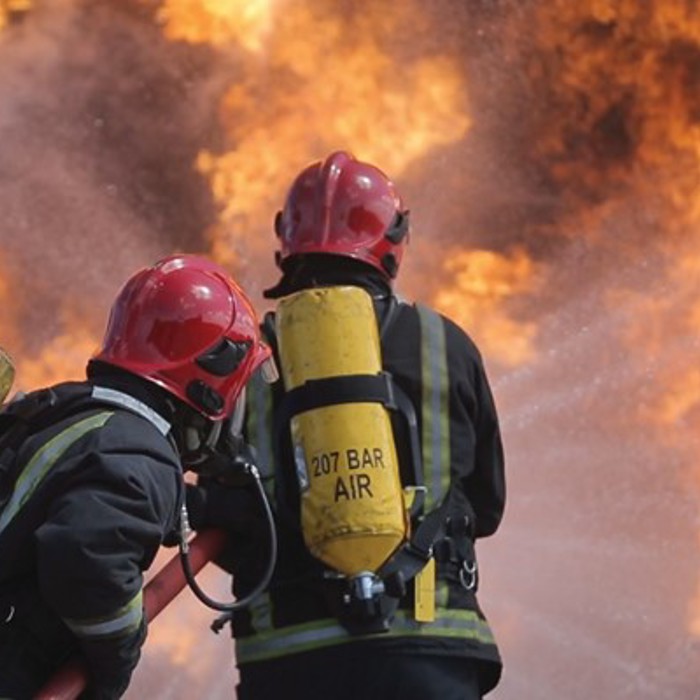 Two firefighters in protective gear and helmets combat intense flames, with water hoses directed at the fire.