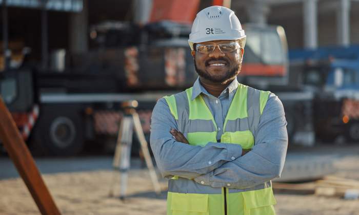 Un trabajador de la construcción con chaleco de seguridad y casco permanece de pie con los brazos cruzados en una obra.