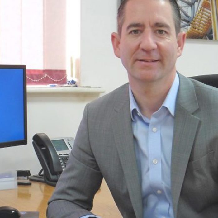 A man in a suit sits at a desk in an office with a computer monitor displaying the text "Drilling Systems" beside him.