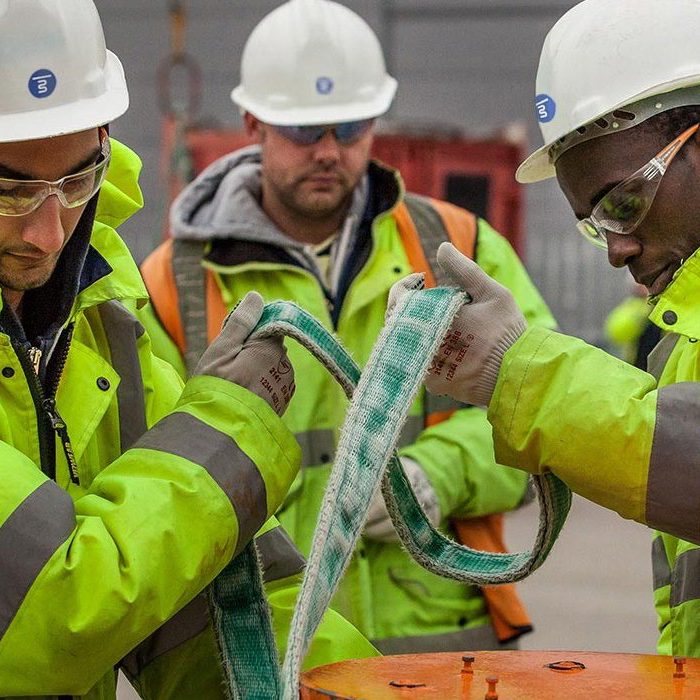 Three construction workers wearing hard hats and high-visibility jackets are securing a strap around an object at a construction site.