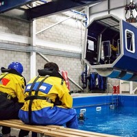 Un grupo de personas vestidas con equipos de seguridad amarillos y azules se sientan en bancos y observan una cabina de helicóptero simulada suspendida sobre una piscina en un centro de formación cubierto.
