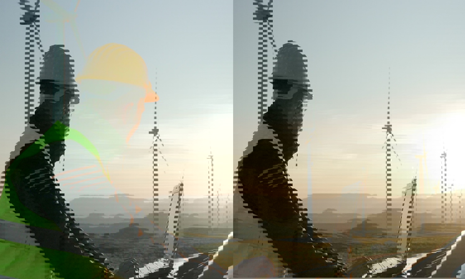 Persona con casco y chaleco de seguridad utilizando un ordenador portátil al aire libre cerca de turbinas eólicas, con un paisaje montañoso de fondo durante la puesta de sol.