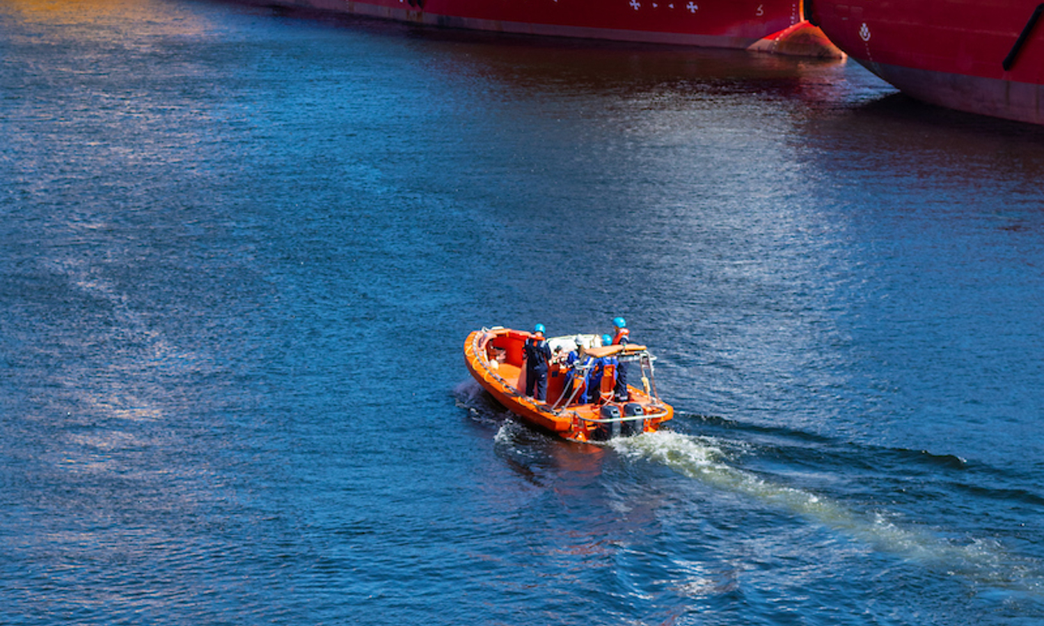 An orange rescue boat with a group of people navigates through blue water, smoothly passing large red ships, showcasing their training and expertise.