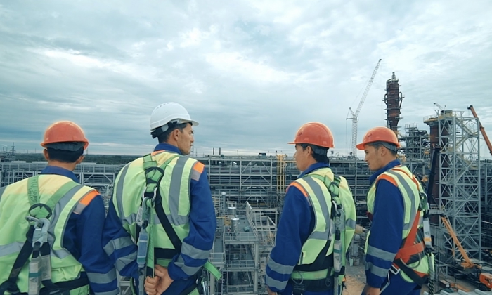 Cuatro trabajadores de la construcción con equipo de seguridad discuten en una obra industrial, con grúas y estructuras de acero visibles al fondo bajo un cielo nublado.