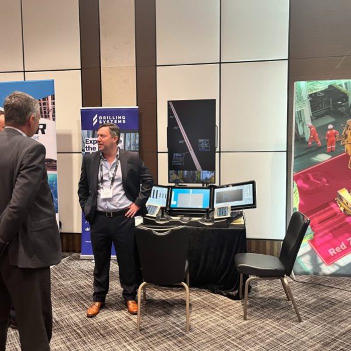 Three men in business attire engage in conversation at a technology expo booth showcasing drilling systems with multiple computer monitors and demonstration banners.