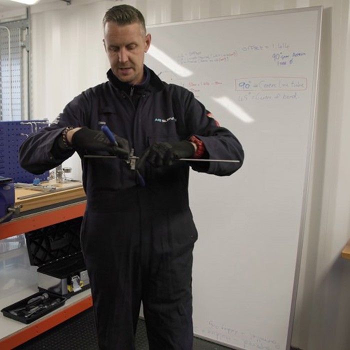 A person in a workshop is using a hand tool to work on a piece of metal, with a whiteboard in the background displaying some notes. Shelving and various tools are visible around the workshop.