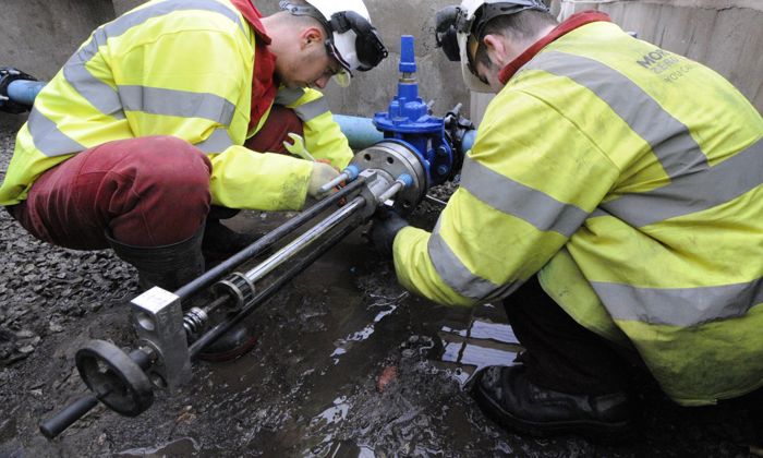 Dos trabajadores con chalecos de seguridad amarillos y cascos están arreglando una válvula de tubería al aire libre sobre un suelo embarrado.