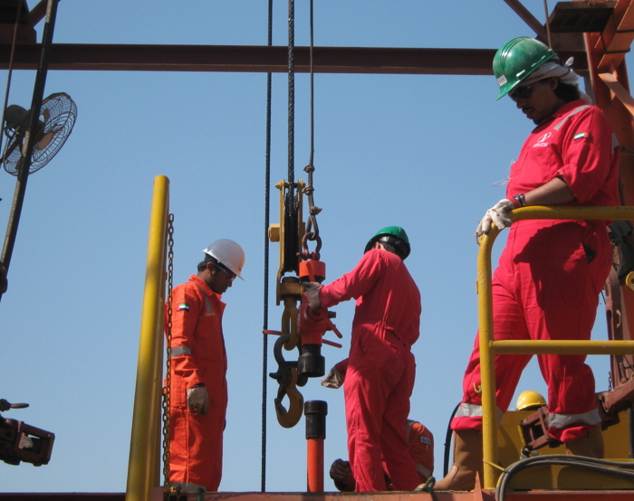Trabajadores con monos y cascos rojos manejan equipos en una plataforma petrolífera bajo un cielo despejado.