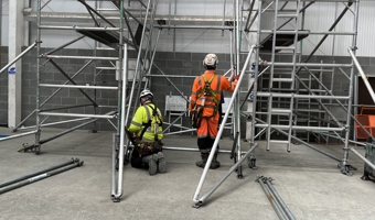 Two workers in safety gear carefully assemble scaffolding in an industrial setting, demonstrating precise training and expertise.