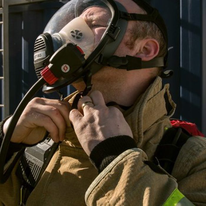 A firefighter adjusts his face mask, wearing protective gear in front of a fire station.