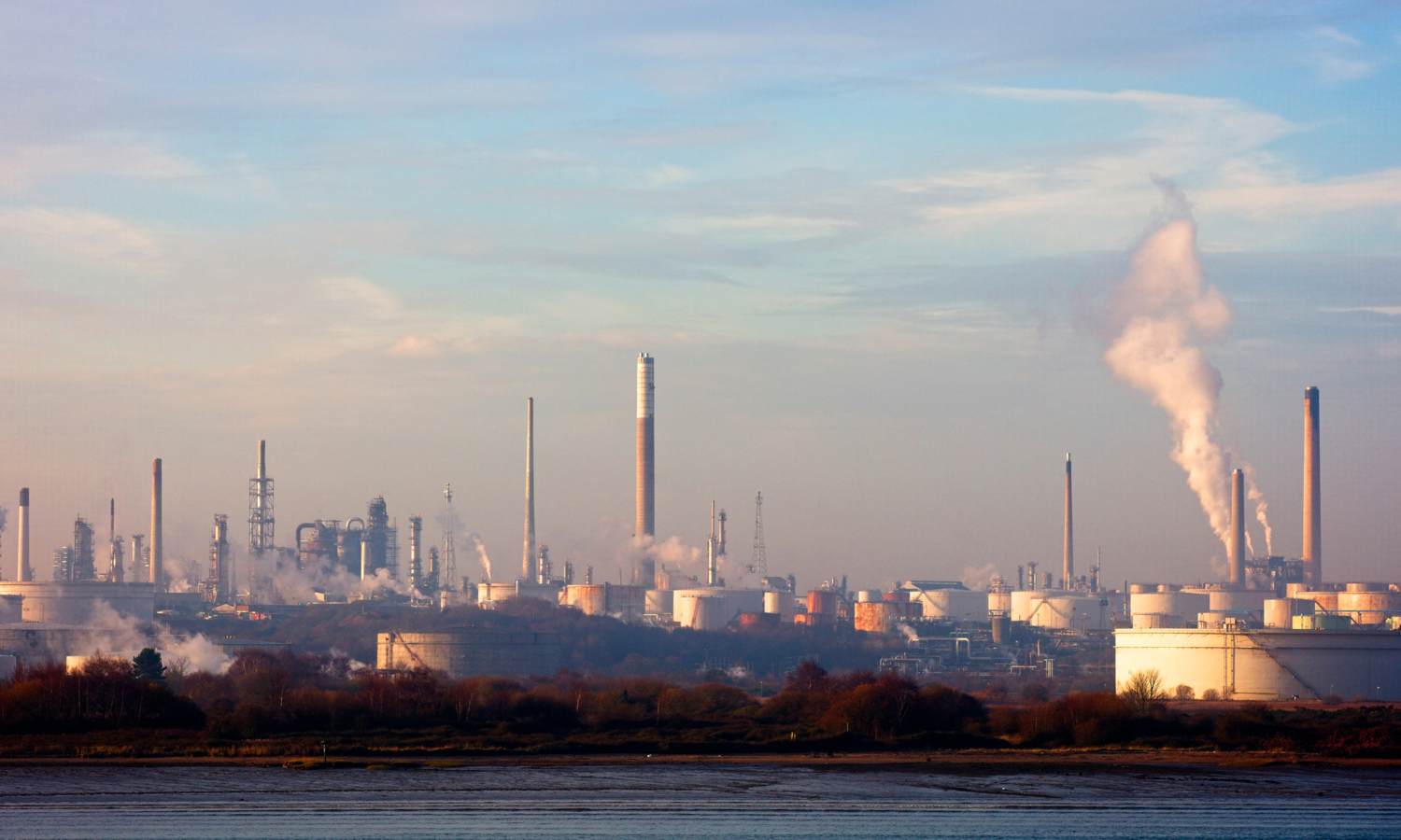 El paisaje de una refinería industrial se extiende bajo un cielo azul con nubes dispersas, sus chimeneas echan humo como centinelas en formación, sincronizadas y firmes.