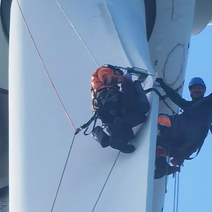 Two workers in safety gear are suspended on the blades of a wind turbine, with one worker waving at the camera.