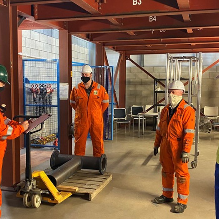 Four workers in safety gear have a discussion in an industrial setting, with materials and equipment visible in the background.