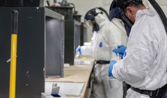 Two people in protective suits and helmets engage in a training session at the lab bench, carefully handling materials with gloves. A box and a yellow pole are visible in the foreground.
