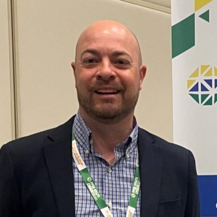 Man in a suit jacket and checkered shirt stands next to a banner reading "FPSO Con".