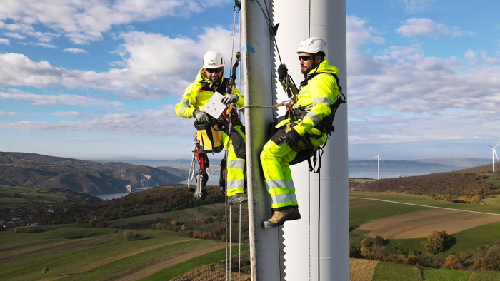 Dos trabajadores con equipo de seguridad, demostrando su formación, inspeccionan un aerogenerador mientras están sujetos con cuerdas, con un paisaje de colinas y más turbinas al fondo.