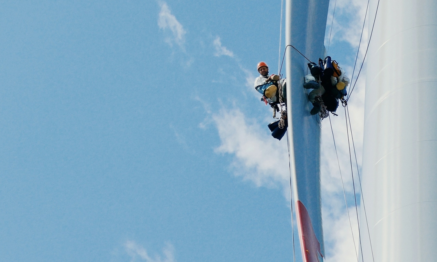 Two workers in safety gear, demonstrating their expert training, are suspended by ropes beside a wind turbine against a blue sky.